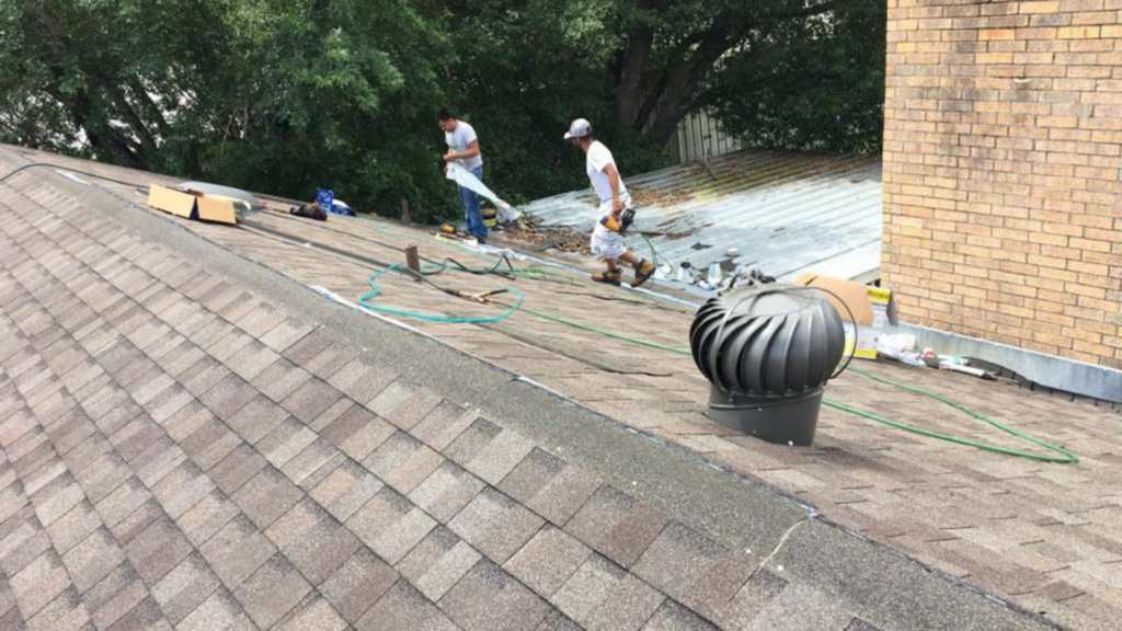 Two workers from a top-rated fencing company repair a rooftop, surrounded by tools and materials. A roof turbine vent is visible in the foreground, with trees and a brick wall in the background.