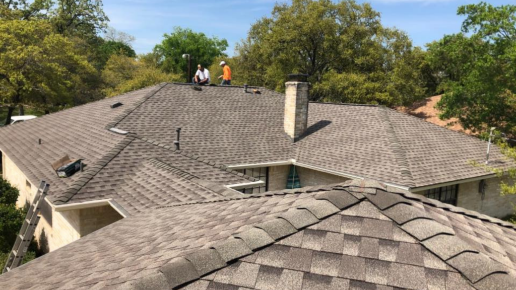 Two workers from a top-rated roofing company are on a large, brown shingled roof with trees in the background and a clear sky.