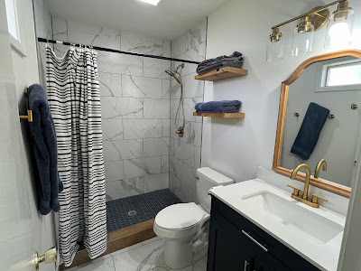 Bathroom remodel near me: A chic space featuring a white tile shower, black and white striped curtain, vanity with a gold faucet, and wooden shelves stacked with plush towels.