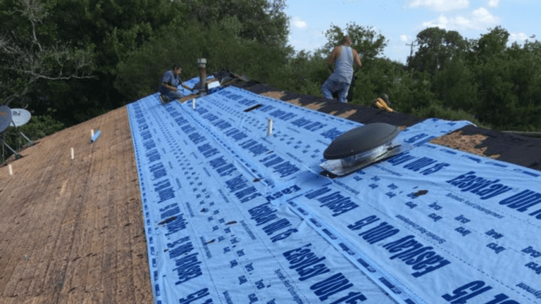 Two workers from a top-rated roofing company install roof underlayment on a wooden roof, surrounded by trees and a satellite dish in the background.