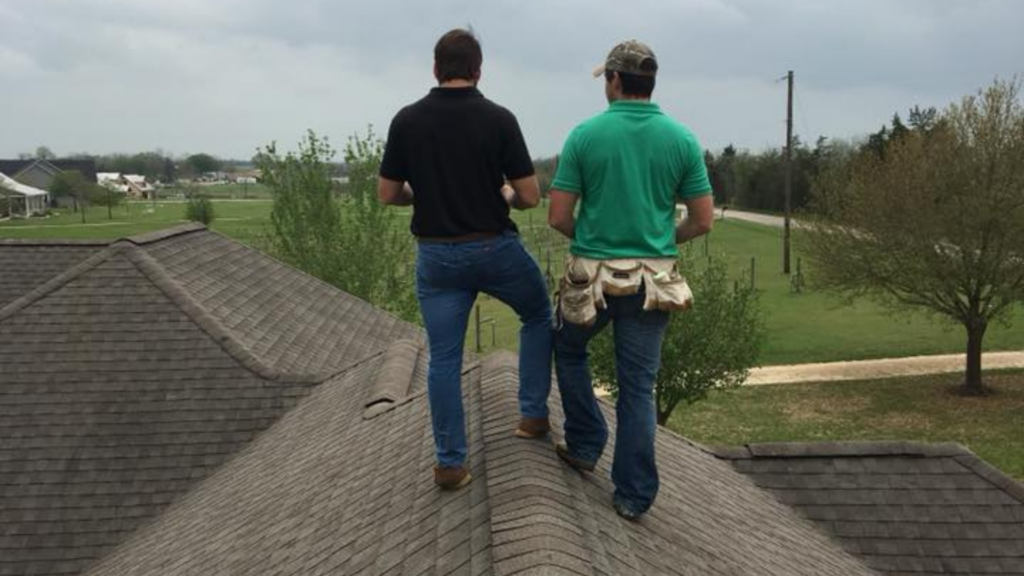 Two people standing on a rooftop, one wearing a black shirt and jeans, the other in a green shirt with a tool belt, likely from a top-rated fencing company. Trees and a road stretch into the distance as they discuss remodeling near me options.