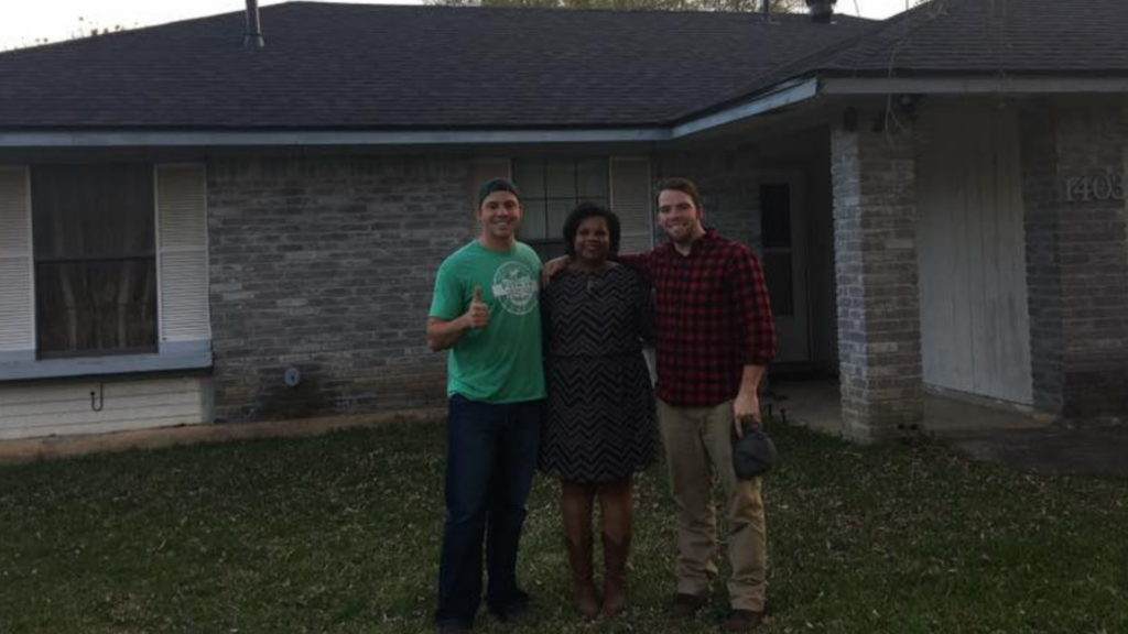 Three people stand outside a brick house with a dark roof, perhaps discussing plans with the top-rated fencing company. The person on the left wears a green shirt, the one in the middle sports a patterned dress, and on the right is someone in a plaid shirt.