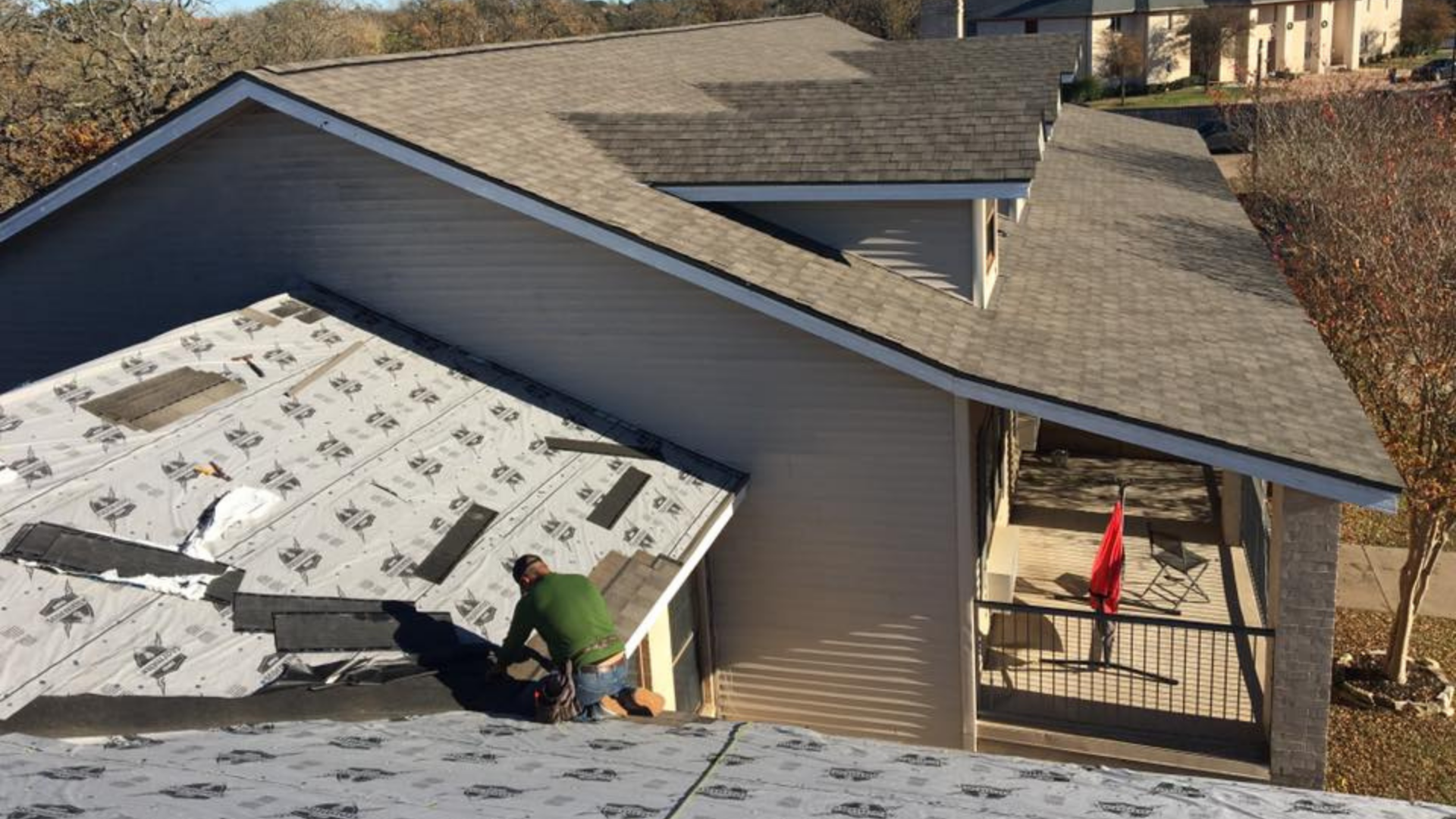 A person is busy installing roofing materials on a sloped roof. While gray shingles cover part of it, another section is laid with underlayment. Nearby, a red flag flutters, marking progress in this roofing near me project.