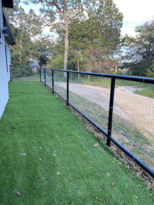 A narrow, grassy yard with a black metal and wire fence runs alongside a white building. Trees and a dirt path lie in the background, nearby one of the top-rated roofing companies known for excellence in remodeling.