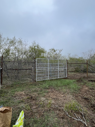 A white metal gate in a wire fence amidst a grassy area with sparse trees under a cloudy sky evokes tranquility. Nearby, where paper bags rustle gently, you might just discover the perfect spot for remodeling near me to transform this serene scene into something extraordinary.