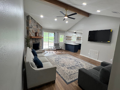 Living room with a stone fireplace, gray sofa, armchair, and patterned rug beneath a ceiling fan. A wall-mounted TV completes the cozy ambiance. The open kitchen features wooden shelves and a black island. Perfect inspiration if youre considering remodeling near me!.