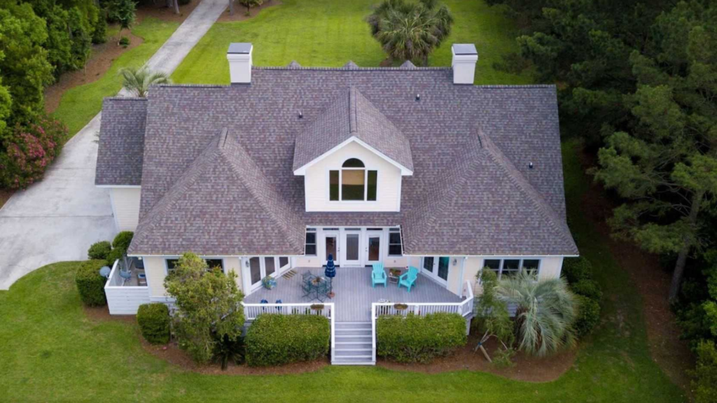 Aerial view of a large house with a gable roof by a top-rated roofing company, surrounded by trees and a lawn. The house features a spacious deck with outdoor furniture and a white railing.