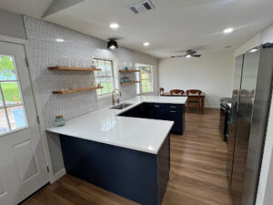 Modern kitchen with wooden floor, white countertops, and dark blue cabinets. Open shelving on tiled wall, stainless steel appliances, and a dining area with wooden furniture in the background. Ideal for those considering remodeling near me to elevate their culinary space.