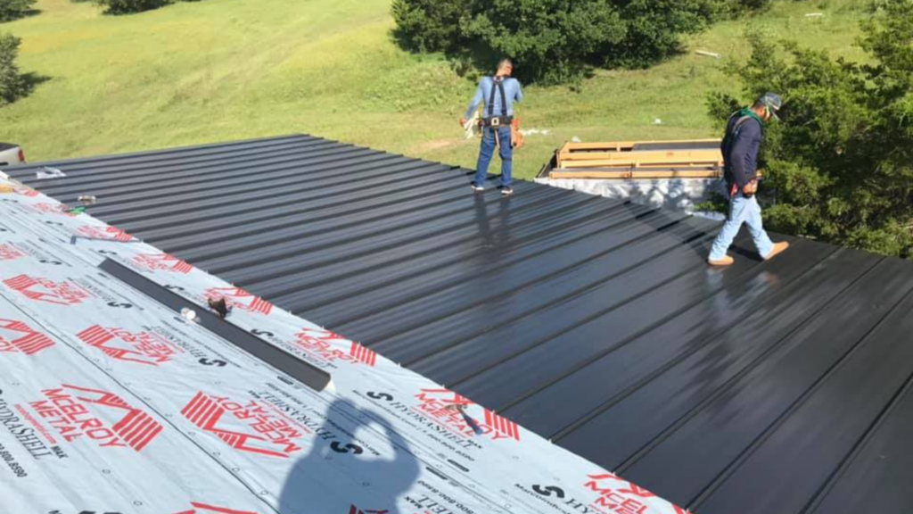 Two workers on a roof under construction, one on metal panels and the other on roofing underlayment, surrounded by greenery—a perfect scene of remodeling near me.