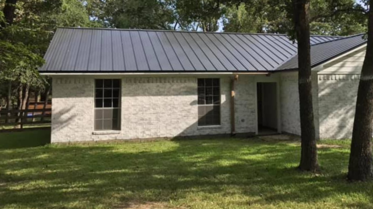 Single-story white brick house with a dark metal roof, two large windows, and a lush grassy lawn surrounded by trees. Ideal for those considering remodeling near me to enhance their outdoor oasis.