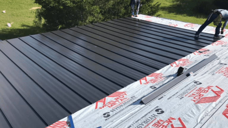 Workers are installing dark metal roofing sheets over insulation material on a building, surrounded by lush greenery. This meticulous project complements ongoing remodeling near me, enhancing both functionality and aesthetic appeal in the neighborhood.