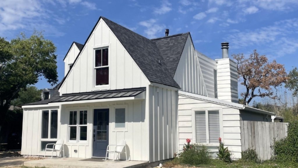 A charming two-story white house with a black roof, two dormer windows, and a chimney stands gracefully amidst the trees under a partly cloudy sky. The front patio, featuring two chairs, invites relaxation. Perfect for those considering remodeling near me to enhance their serene space.