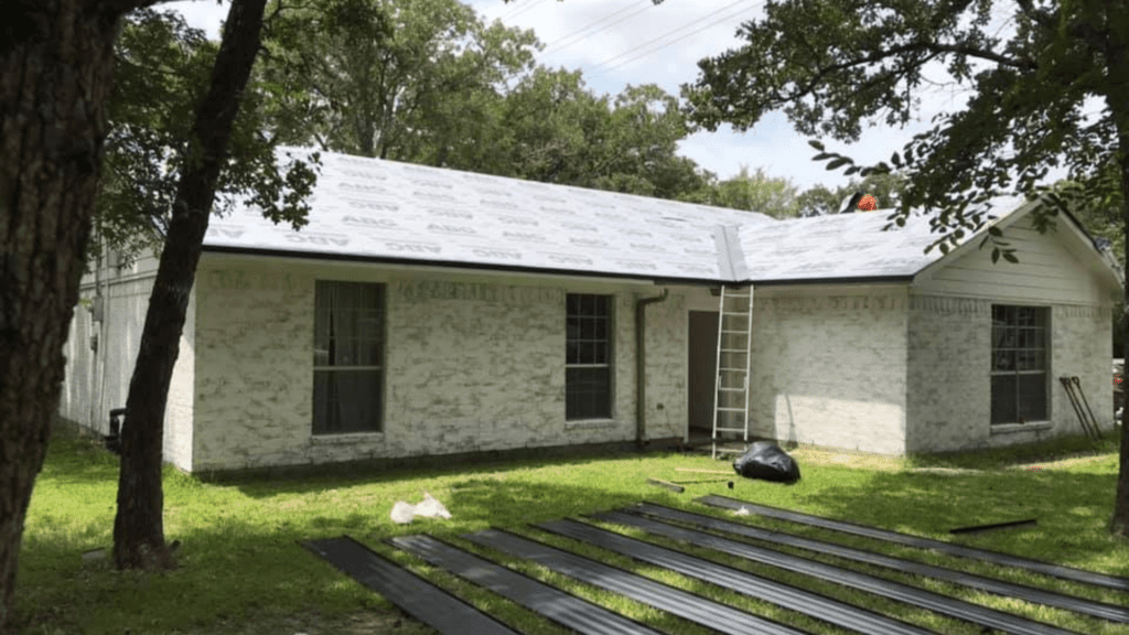 A house with a white exterior is under construction, featuring roofing material laid on the roof and metal sheets scattered nearby. A ladder leans against it as trees surround the area, providing a picturesque backdrop for this remodeling near me project.