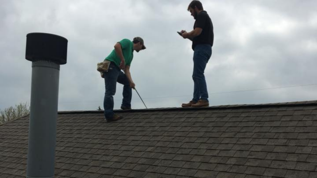 Two men standing on a sloped roof; one is inspecting with a tool, while the other holds a device, likely planning updates. Overcast sky looms as they focus on their task. Nearby, the top-rated fencing company prepares for an upcoming project.