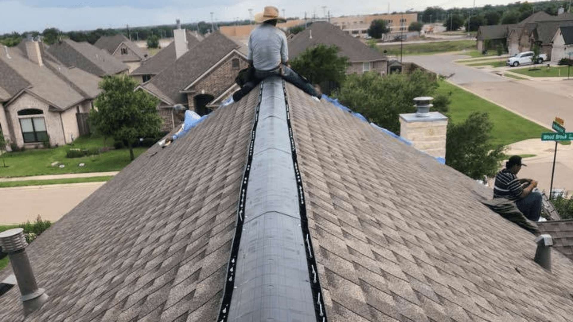 Two people sit on a peaked residential roof. One person wears a straw hat, perhaps considering options for remodeling near me. Houses and green lawns stretch out in the background under a cloudy sky.