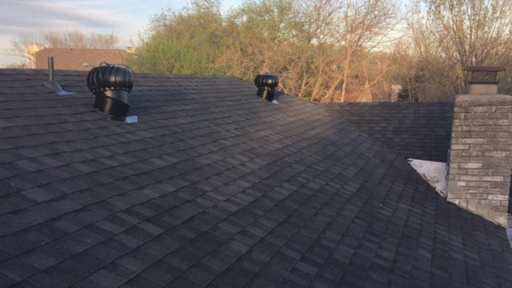 A rooftop with dark shingles, showcasing two turbine vents and a chimney, reflects a recent remodeling near me. In the backdrop, trees and a house complete the view.