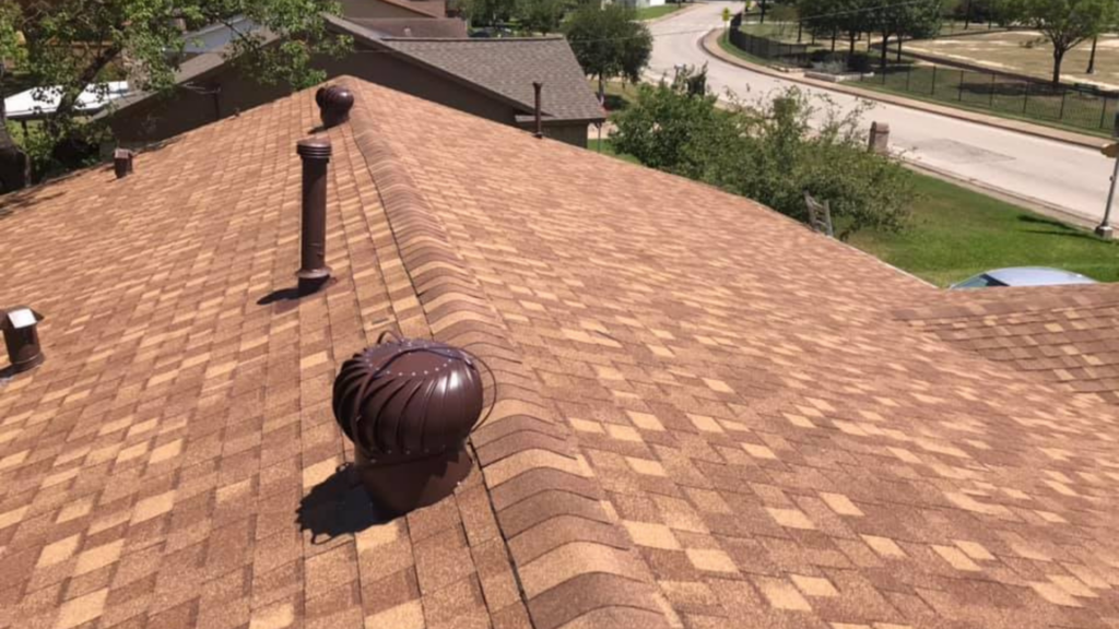 Brown shingled roof with a vent and wind turbine vent, nestled amidst trees and a road. This home benefits from the expertise of a top-rated roofing company, ensuring durability and peace of mind.