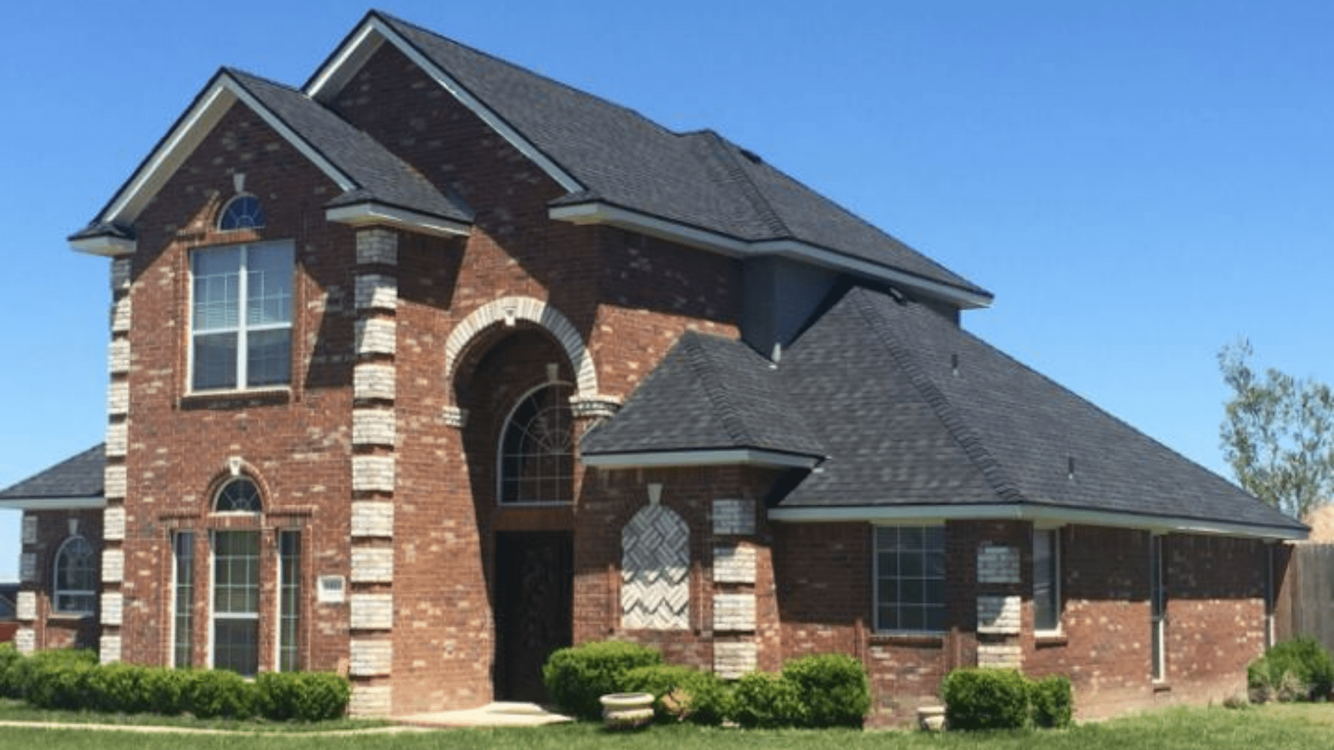 A two-story brick house with black roof shingles features large arched windows, surrounded by small green bushes against a clear blue sky. Nearby, a top-rated fencing company enhances the landscape with elegantly designed enclosures that beautifully complement the homes classic architecture.