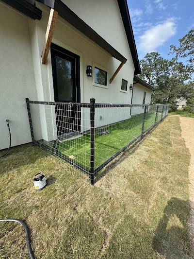 A fenced-off grassy area stretches beside a white house with a black door. Outside the fence, a canister and shadows lie on the grass—perfect for those considering remodeling near me to enhance their outdoor space.