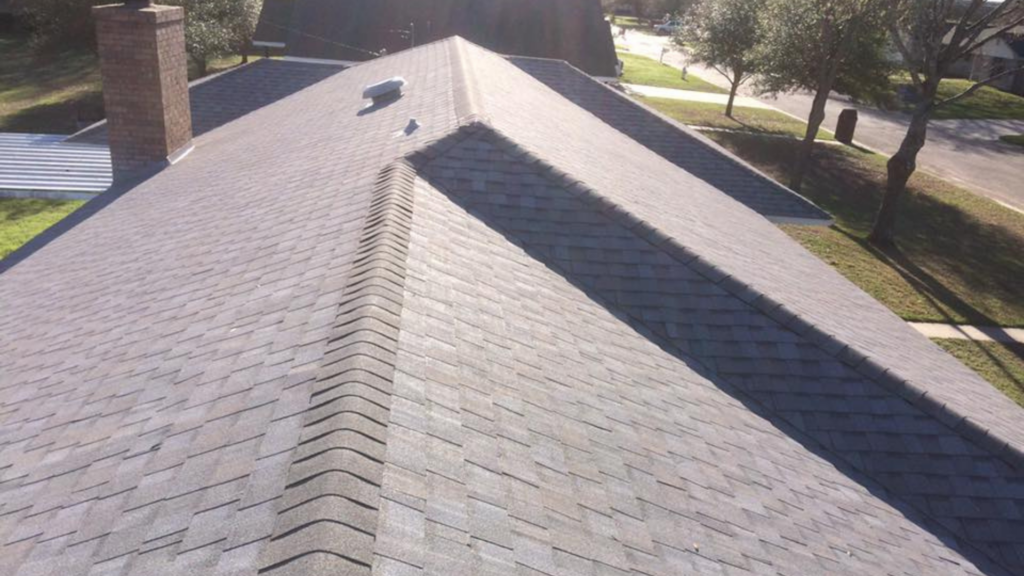 A shingled residential roof with a chimney and vent is shown, epitomizing quality roofing near me. The sun casts shadows, highlighting the roofs peak and angles. Trees and a street are visible in the background, emphasizing the perfect blend of nature and design.